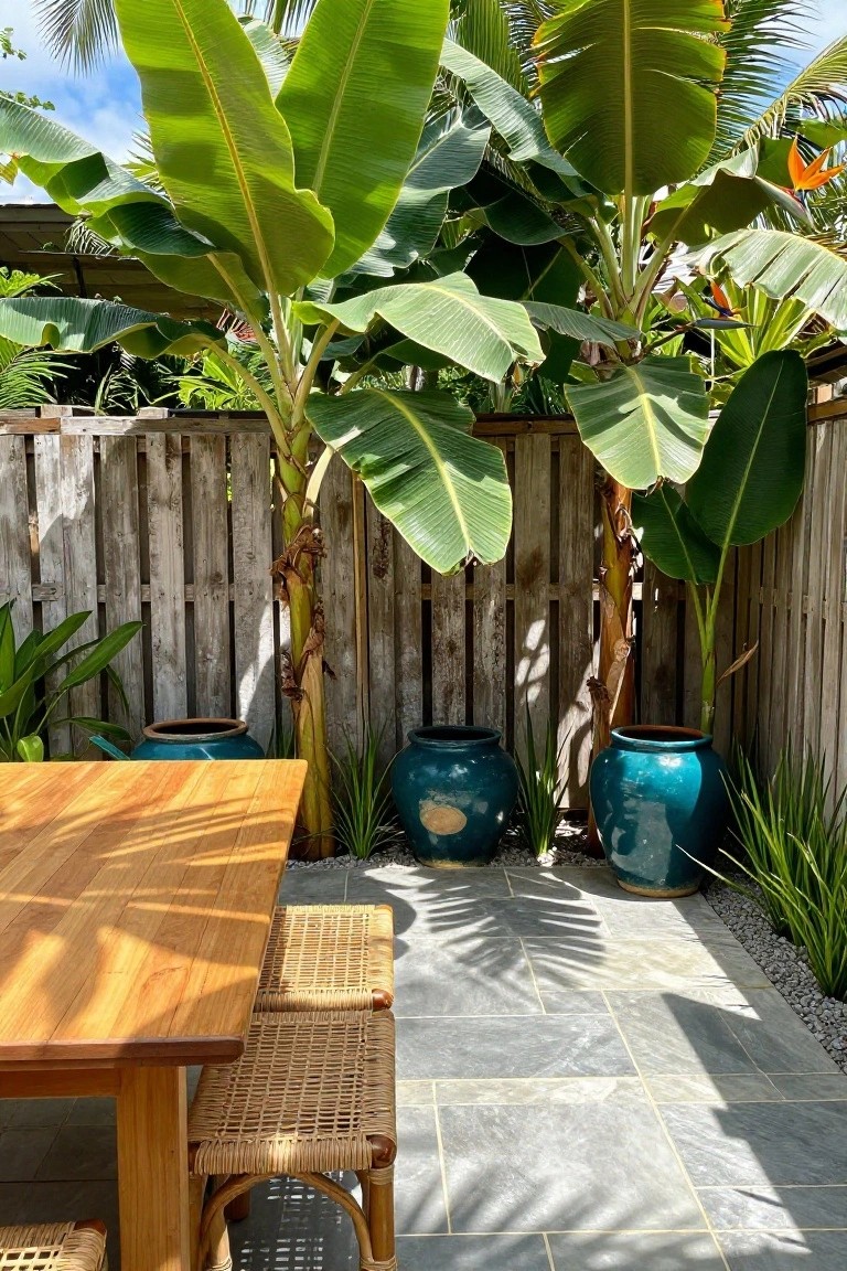 Weathered vertical wood pallet fence enclosing a backyard patio with a wooden table, rattan stools, large turquoise ceramic pots containing grasses, banana trees, and other tropical plants on a slate tile floor with gravel edging.