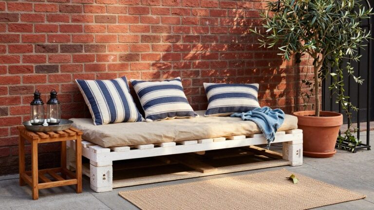 Whitewashed wooden pallet sofa with beige cushions, blue-and-white striped pillows, and blue throw blanket on a paved outdoor patio against a brick wall, with potted olive tree, climbing plants, wooden side table holding glass jars, and seagrass rug.