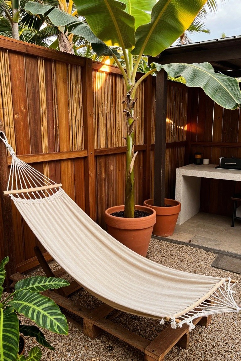 Backyard outdoor space featuring a beige hammock on a wooden pallet stand, enclosed by tall wooden slat and bamboo fence, with banana plants in terracotta pots, gravel flooring, and a concrete counter with stools nearby.
