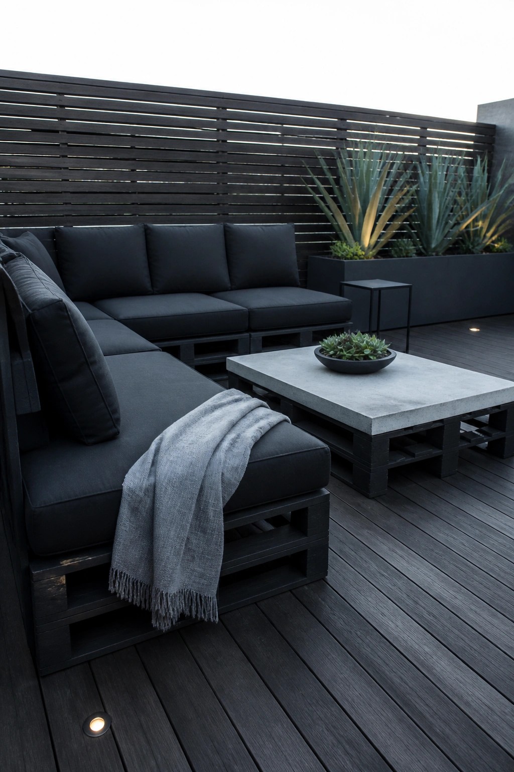 Modern dark pallet sofa with black cushions, gray throw, and concrete table on deck.