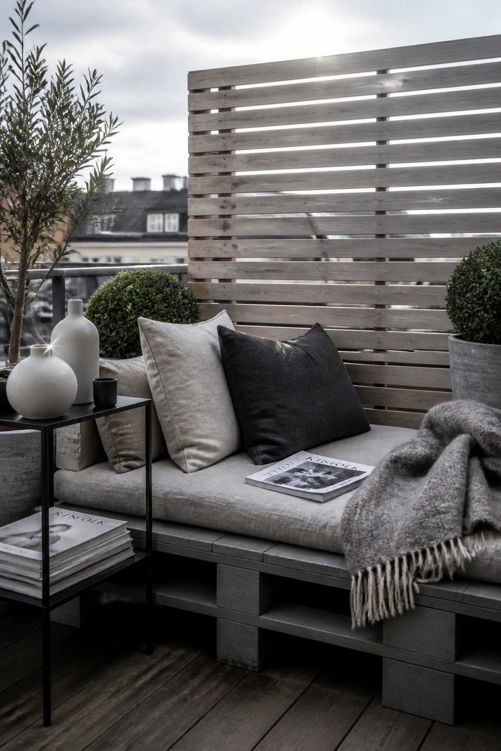An outdoor daybed made from gray wooden pallets on a balcony, with gray cushions, pillows, a fringed throw blanket, and side tables holding magazines and vases.