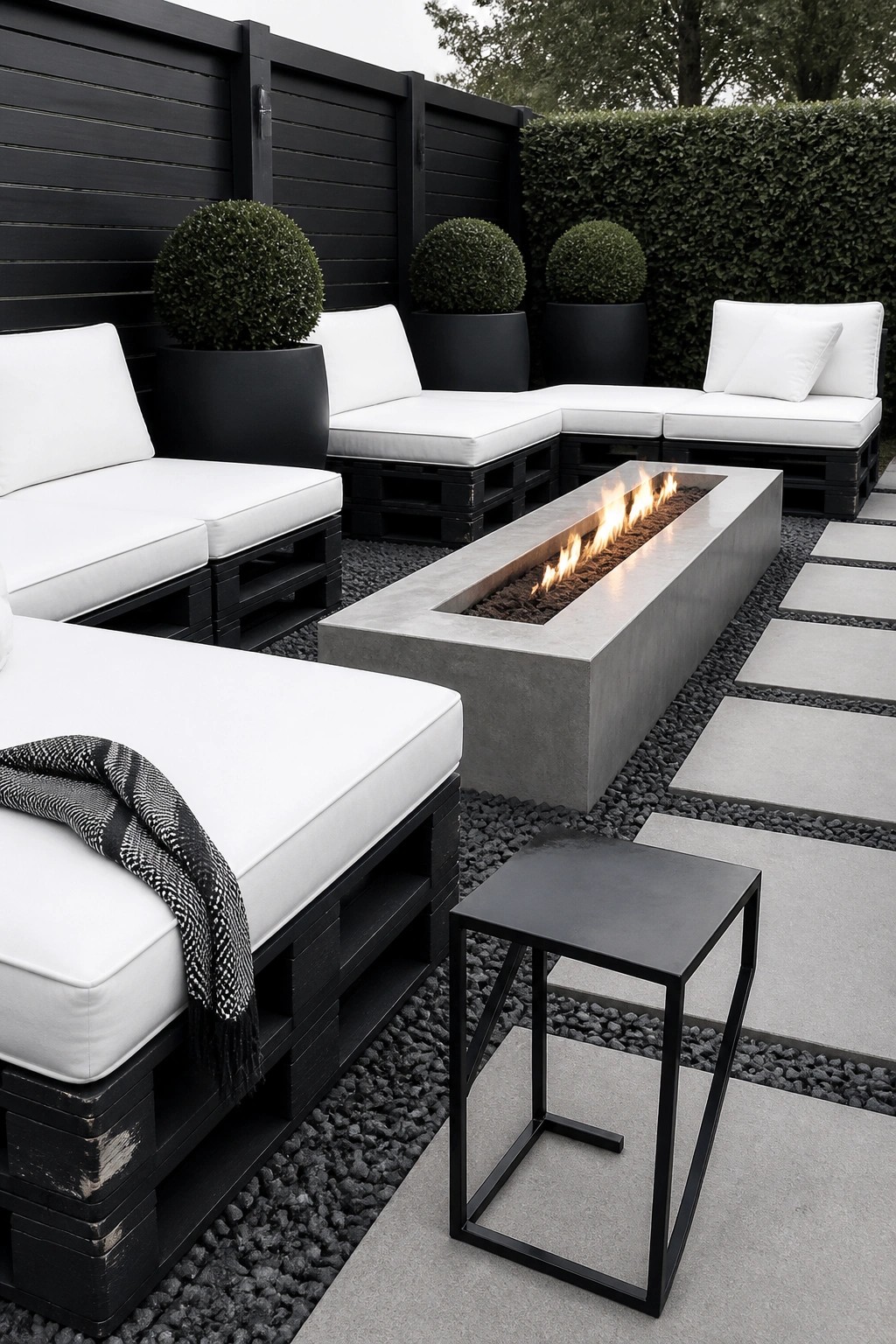 Outdoor patio with black painted pallet sofas with white cushions arranged around a rectangular concrete fire pit on gravel and stone pavers.
