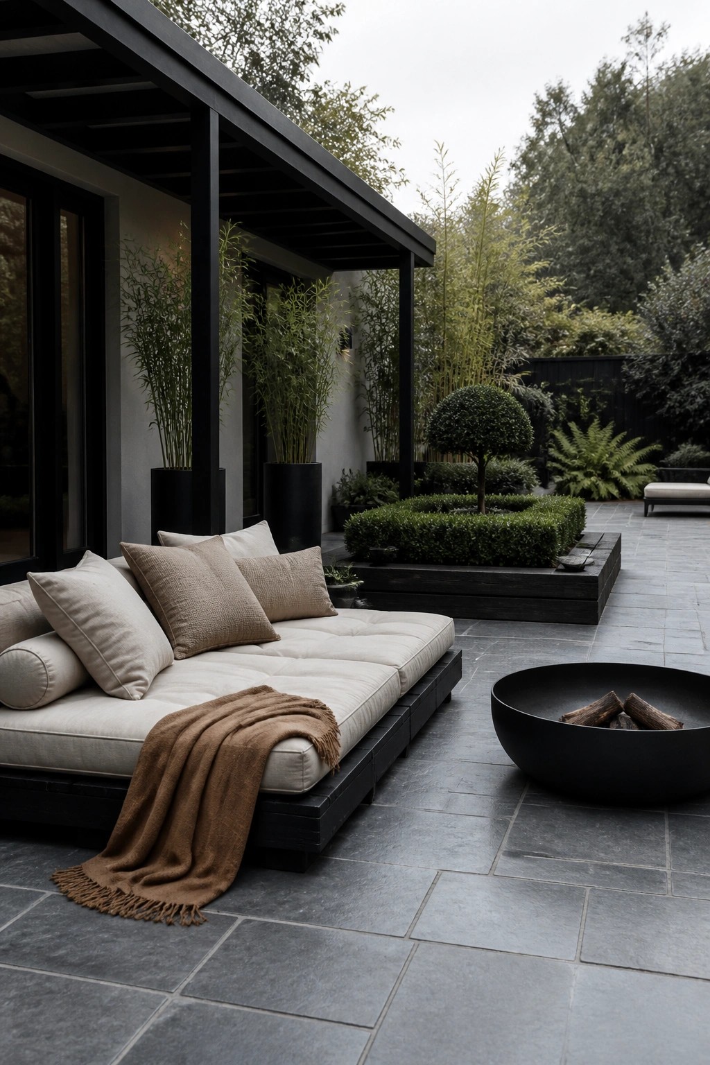 A dark wooden daybed with beige cushions and a brown throw sits on a stone patio beside a round black fire pit, with potted plants and hedges in the background.