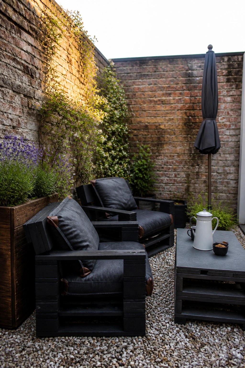 Black painted pallet armchairs with gray cushions sit on gravel beside a low pallet table holding a white teapot, near a brick wall with plants.