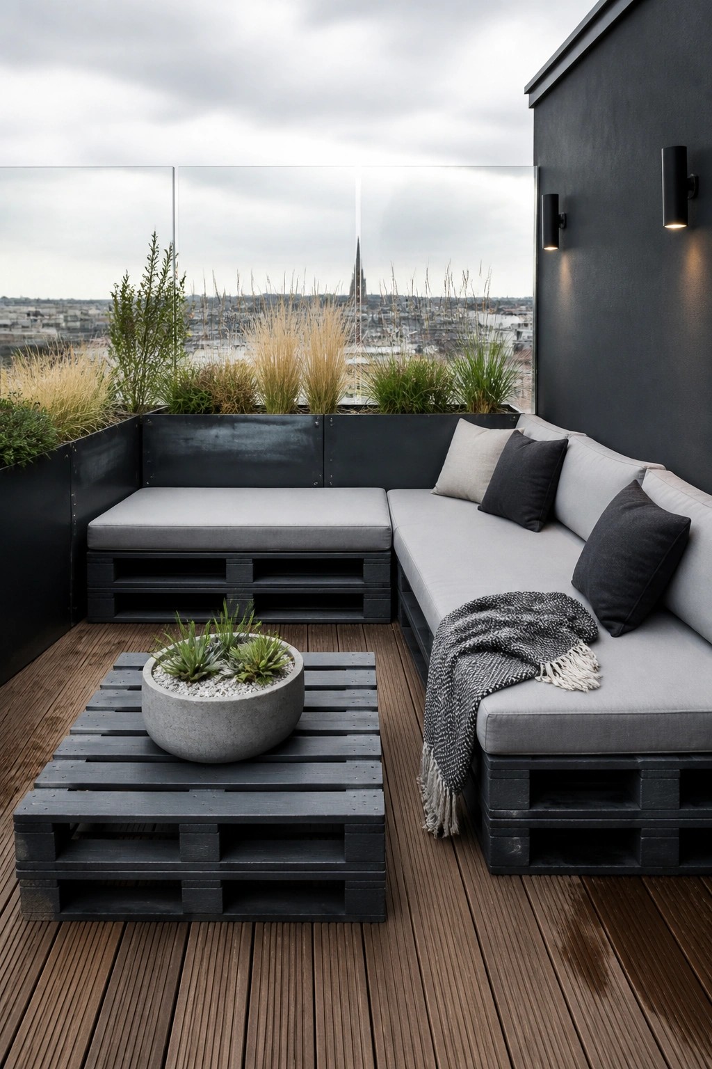 A rooftop terrace featuring L-shaped seating and a low table built from black painted pallets, topped with gray cushions and a concrete planter.