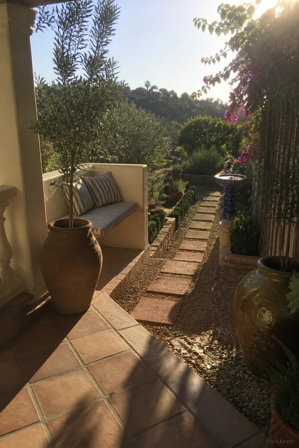 A terracotta-tiled patio with a built-in bench and large clay pots leads to a gravel path of rectangular stepping stones that continues into a landscaped garden with shrubs and trees.