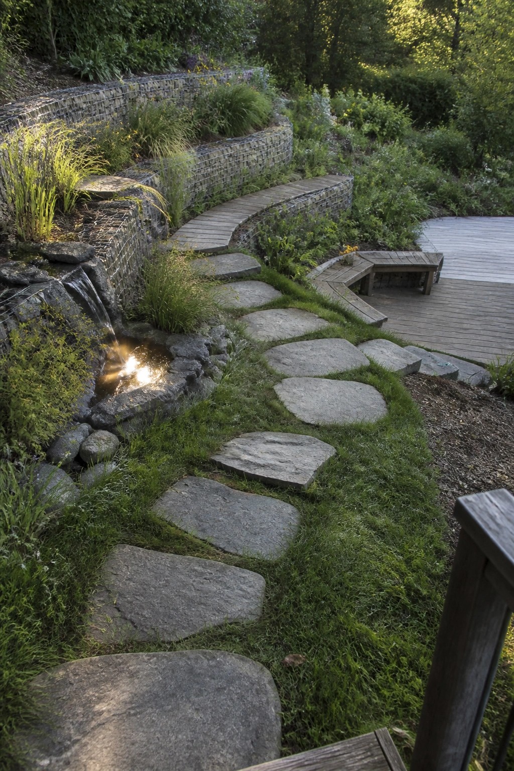 A garden path of large flat stepping stones set into grass, leading past a small waterfall and gabion walls toward a wooden deck.