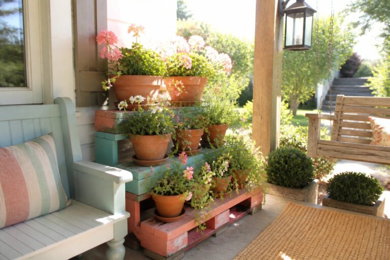 18 Colorful Pallet Plant Stand Ideas for Tiered Flower Displays A multi-tiered wooden plant stand painted orange and teal holds several terracotta pots of pink and white flowers on a wooden porch beside a light blue bench.
