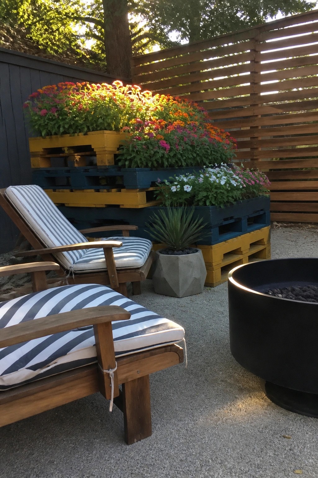 Outdoor patio with striped lounge chairs, a black fire pit, and stacked yellow and blue wooden pallets filled with colorful flowers.