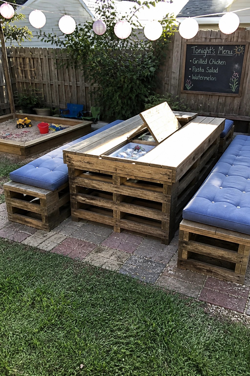 Wooden pallet dining table with hinged lid open over an ice-filled compartment, blue cushioned benches on each side, on a brick patio with string lights overhead.