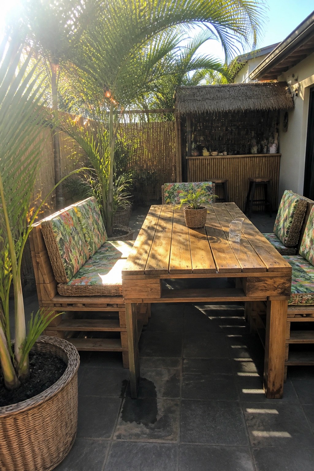 A wooden pallet dining table with matching benches on a stone patio, next to a thatched outdoor bar.