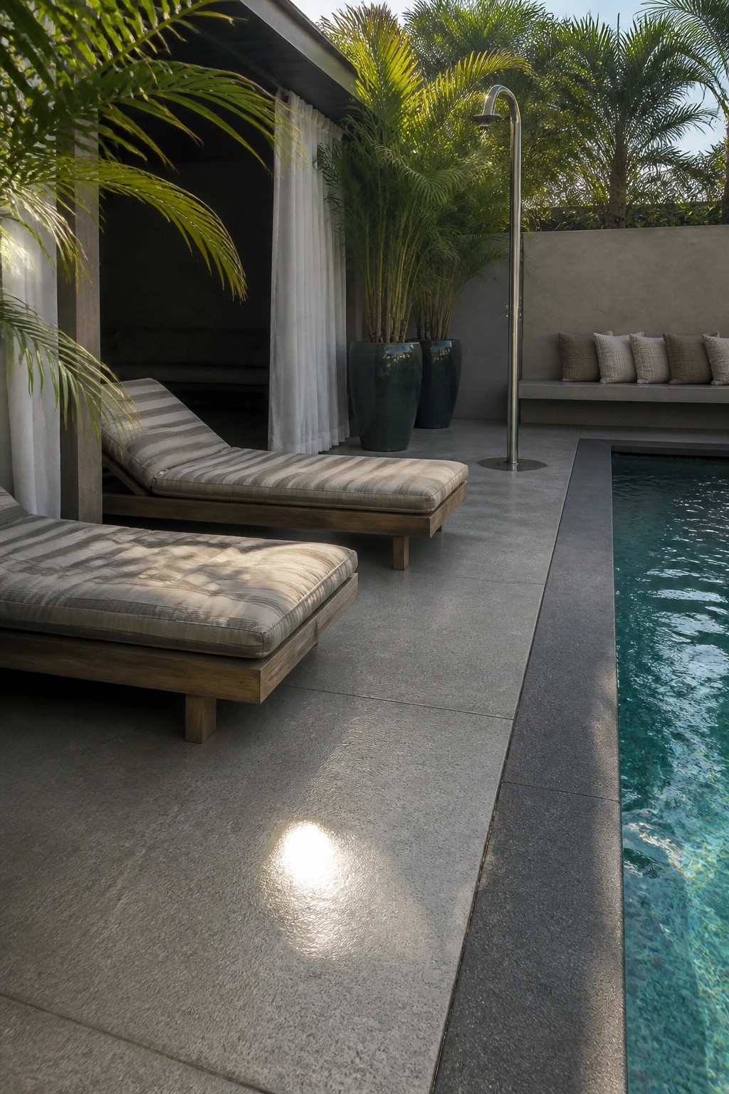 Outdoor patio with large gray concrete slabs beside a swimming pool, two striped lounge chairs, and large potted plants.