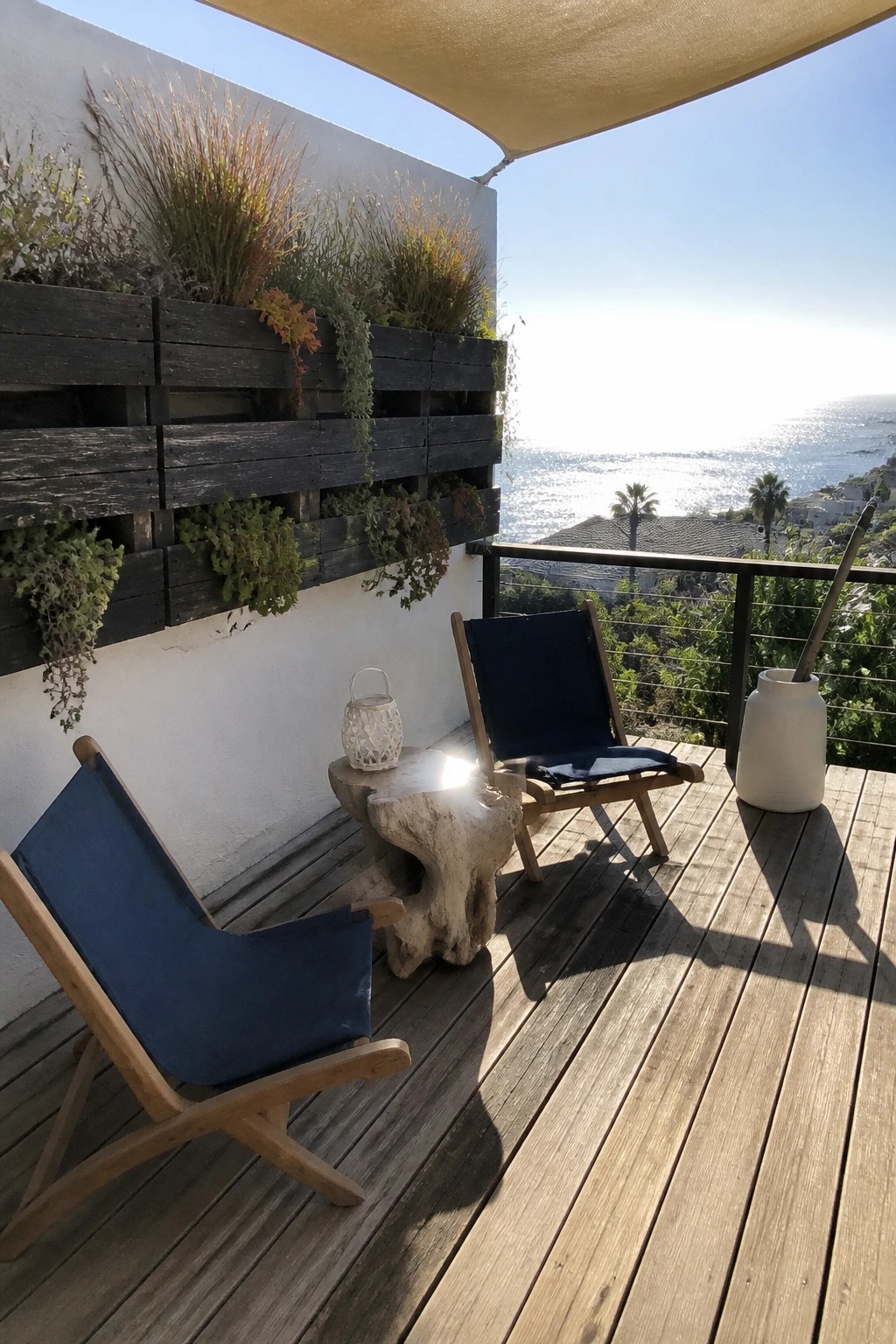 A wooden deck with two blue chairs and a dark stained pallet planter mounted on the white wall, overlooking the ocean.