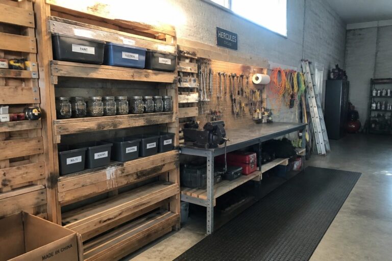 18 Practical Pallet Shelving Ideas for Garage Wall Storage A garage interior with wooden shelves on metal racks holding black labeled storage bins, glass jars, and tools above a workbench.