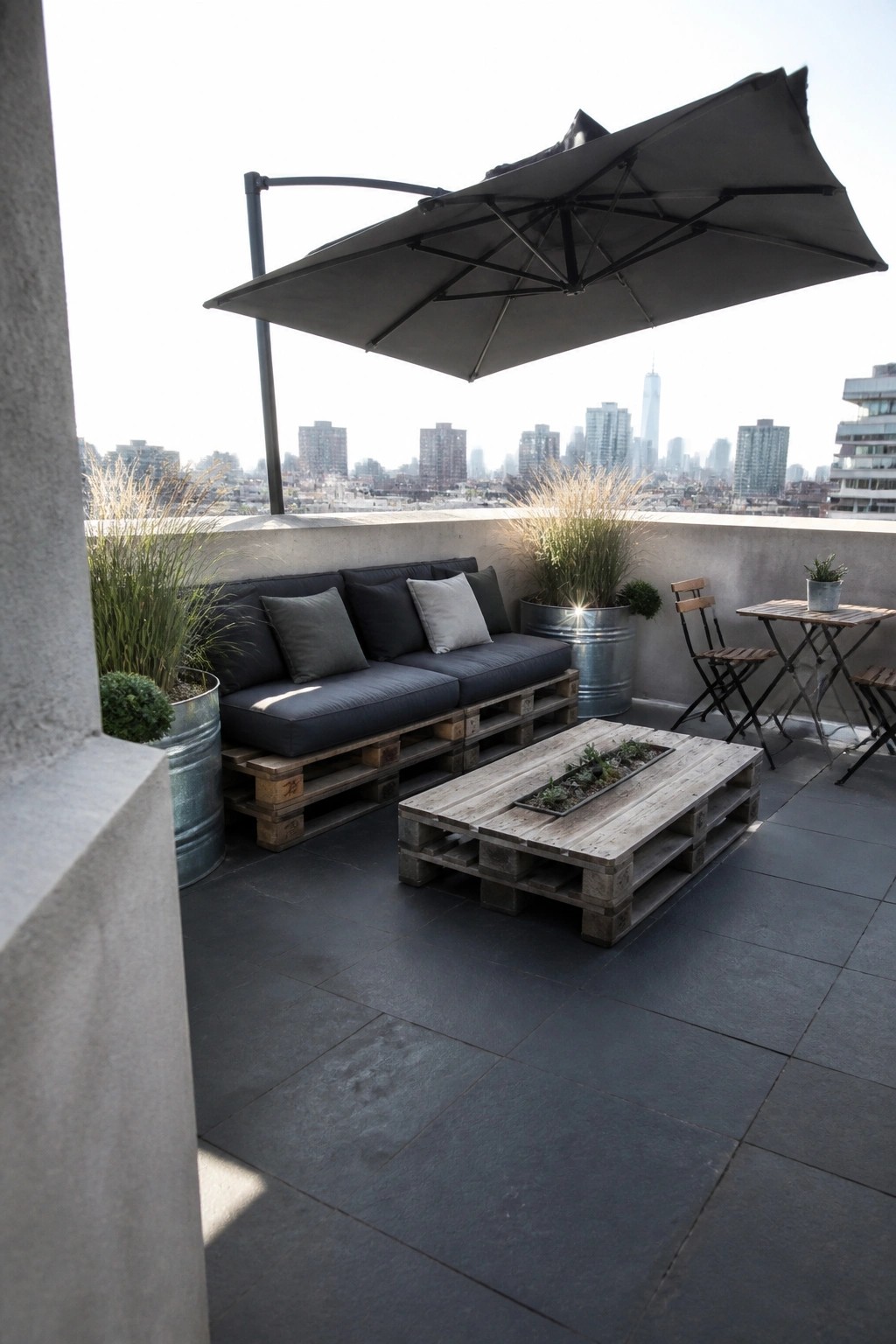 Rooftop patio featuring a pallet sofa with dark cushions, a pallet coffee table, a large cantilever umbrella, and potted plants against a city skyline.