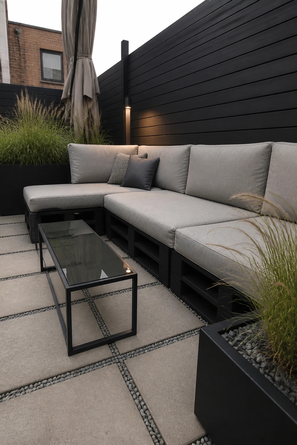 An outdoor patio with an L-shaped sectional sofa made from black painted pallets and gray cushions, placed on large gray tiles with pebble joints and flanked by black planters with tall grasses.