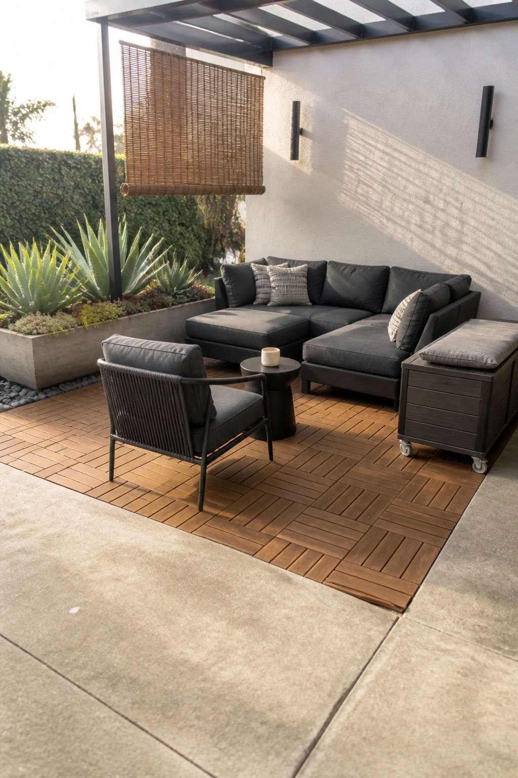 Outdoor patio covered with square brown wooden deck tiles, featuring a dark sectional sofa, black woven armchair, and concrete planter in the background.