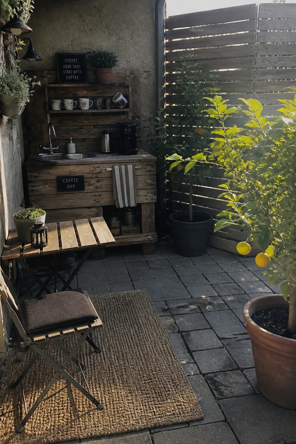 Outdoor pallet coffee bar on a stone patio with sink, shelves holding mugs, coffee maker, striped towel, small folding table and chair on a woven rug, and potted plants nearby.