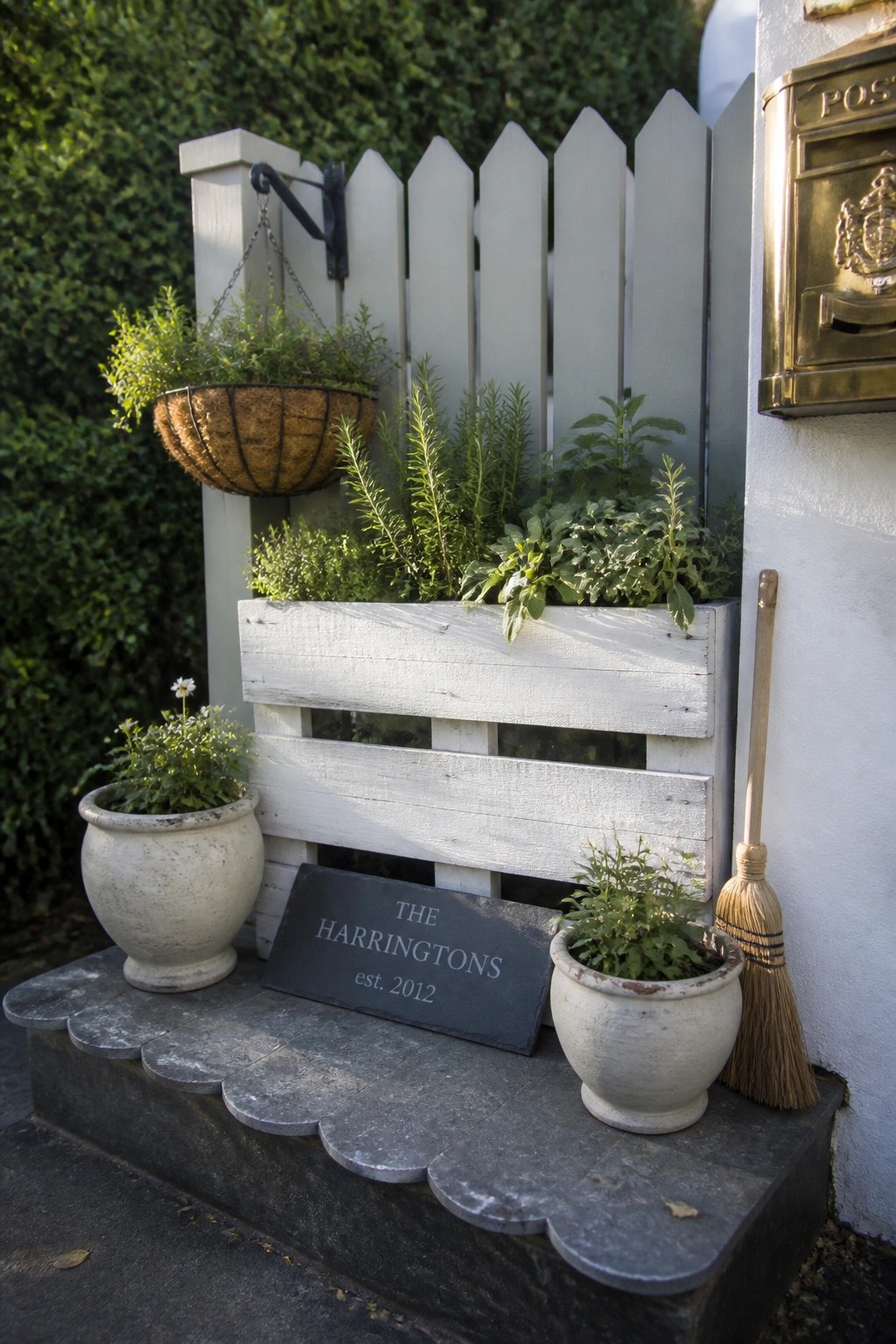 A white painted pallet planter mounted on a fence holds green plants, with potted plants, a hanging basket, and a slate sign on stone steps nearby.