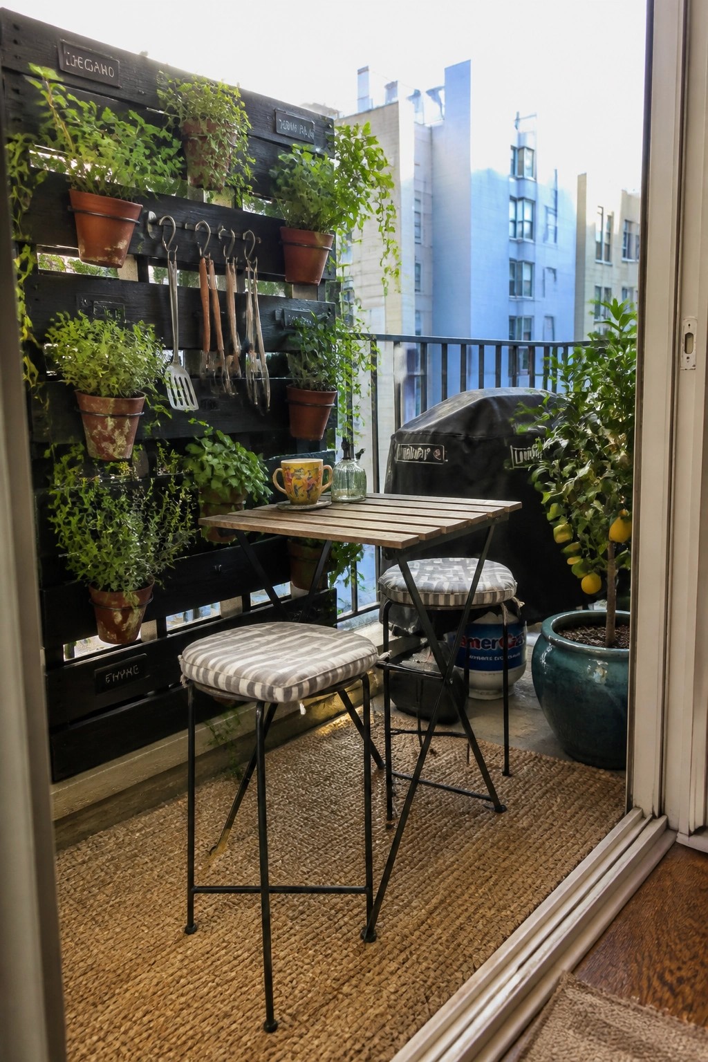 A wooden pallet is mounted vertically on a balcony wall and holds multiple potted plants with gardening tools hanging from hooks.