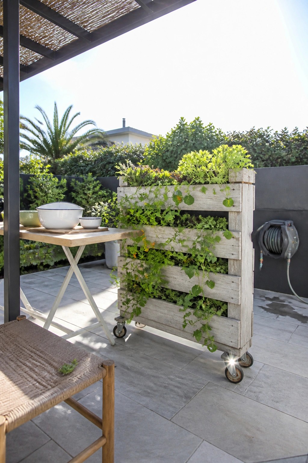 A wooden pallet planter mounted on casters and filled with leafy plants sits on a tiled outdoor patio.
