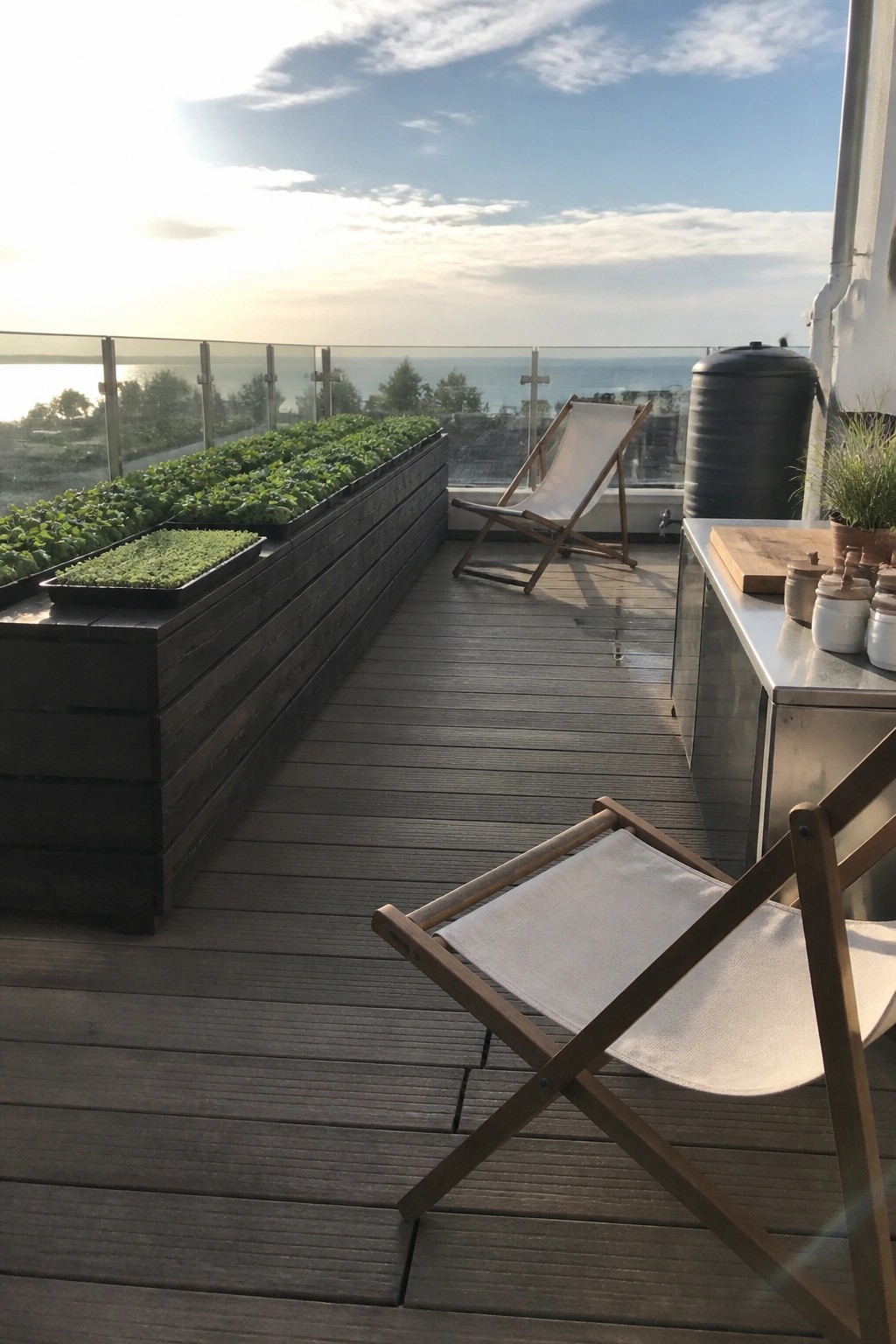 Long dark wooden raised planters filled with green leafy plants sit along the edge of a wooden deck with a glass railing and ocean view.