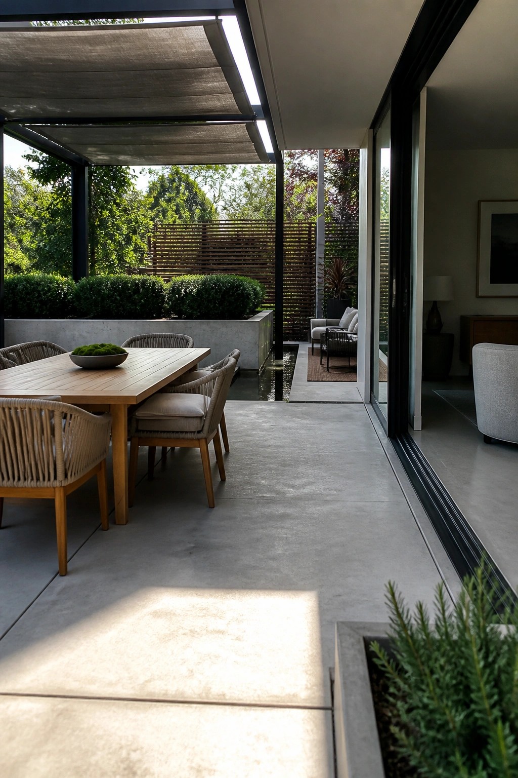 A wooden outdoor dining table with woven chairs sits on a concrete patio beneath a shaded structure beside wide open glass doors leading into a modern home.