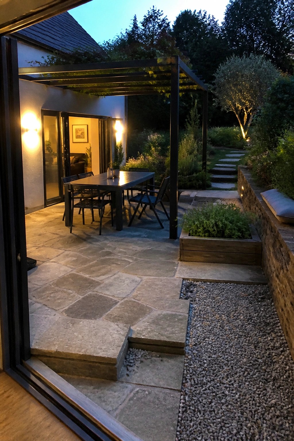 Twilight patio dining area with table under lit pergola beside garden path and stone wall.
