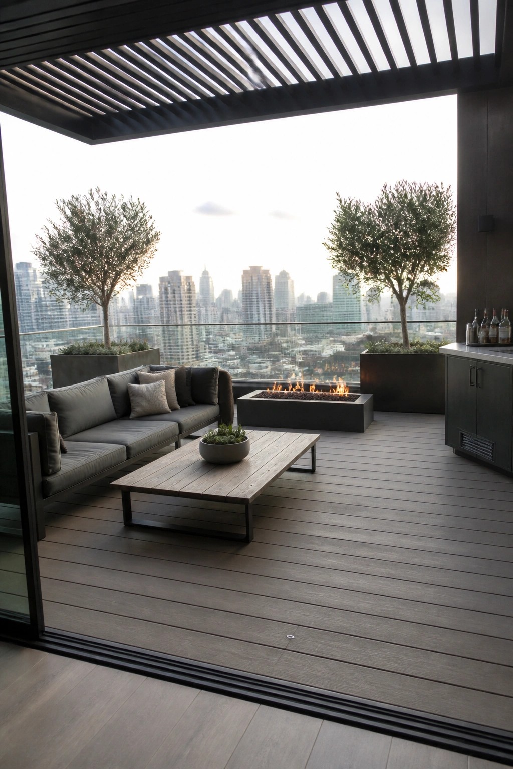 Modern rooftop patio with gray sectional sofa, wooden coffee table, rectangular fire pit, and potted trees, viewed through open sliding glass doors with a city skyline in the background.