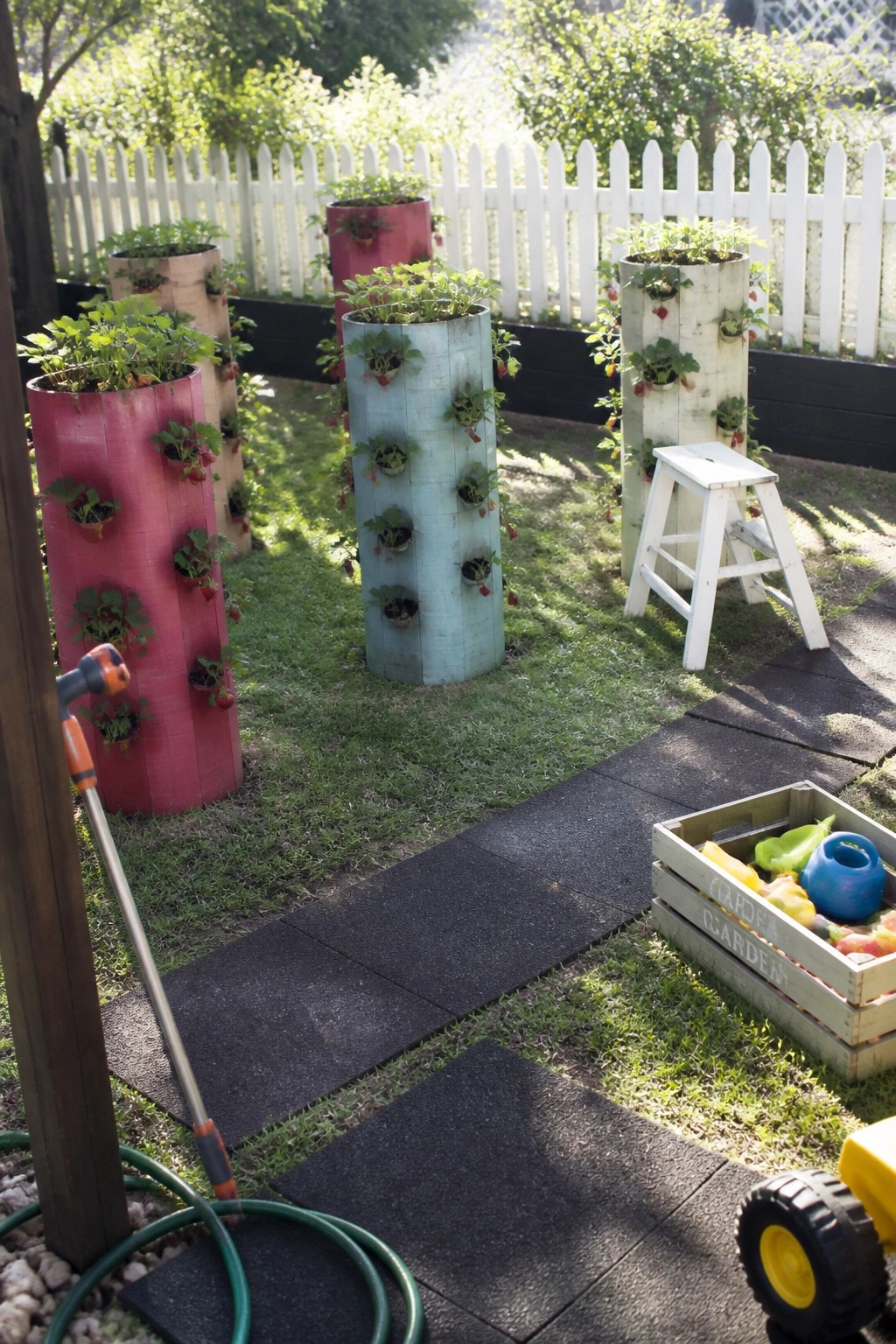 A backyard garden with several tall painted cylindrical planters holding strawberry plants, set on grass beside a dark path with a white stool and wooden crate nearby.