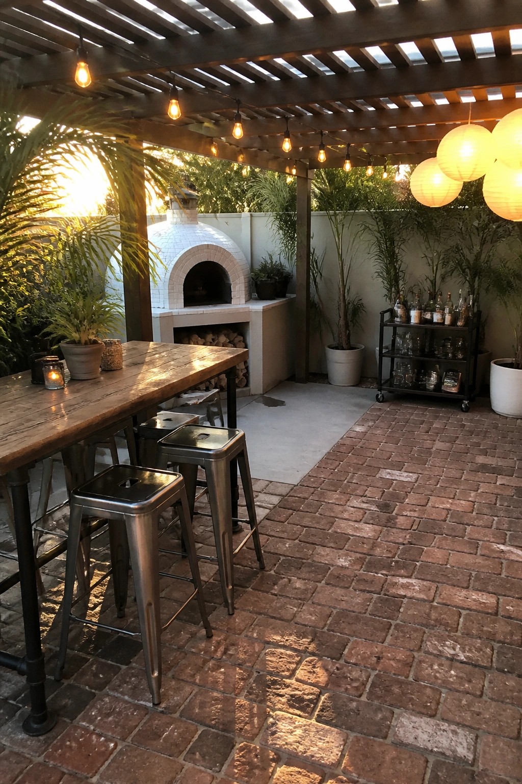 Outdoor patio with a wooden pergola, string lights overhead, a long table with metal stools, a white pizza oven, and potted plants on a brick floor.