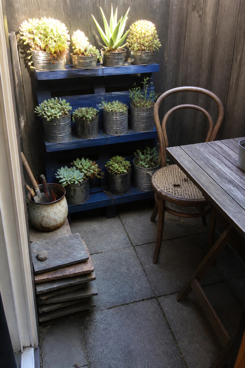 Blue painted wooden pallet used as tiered shelves holding metal buckets of succulents on a stone patio beside a wooden table and chair.
