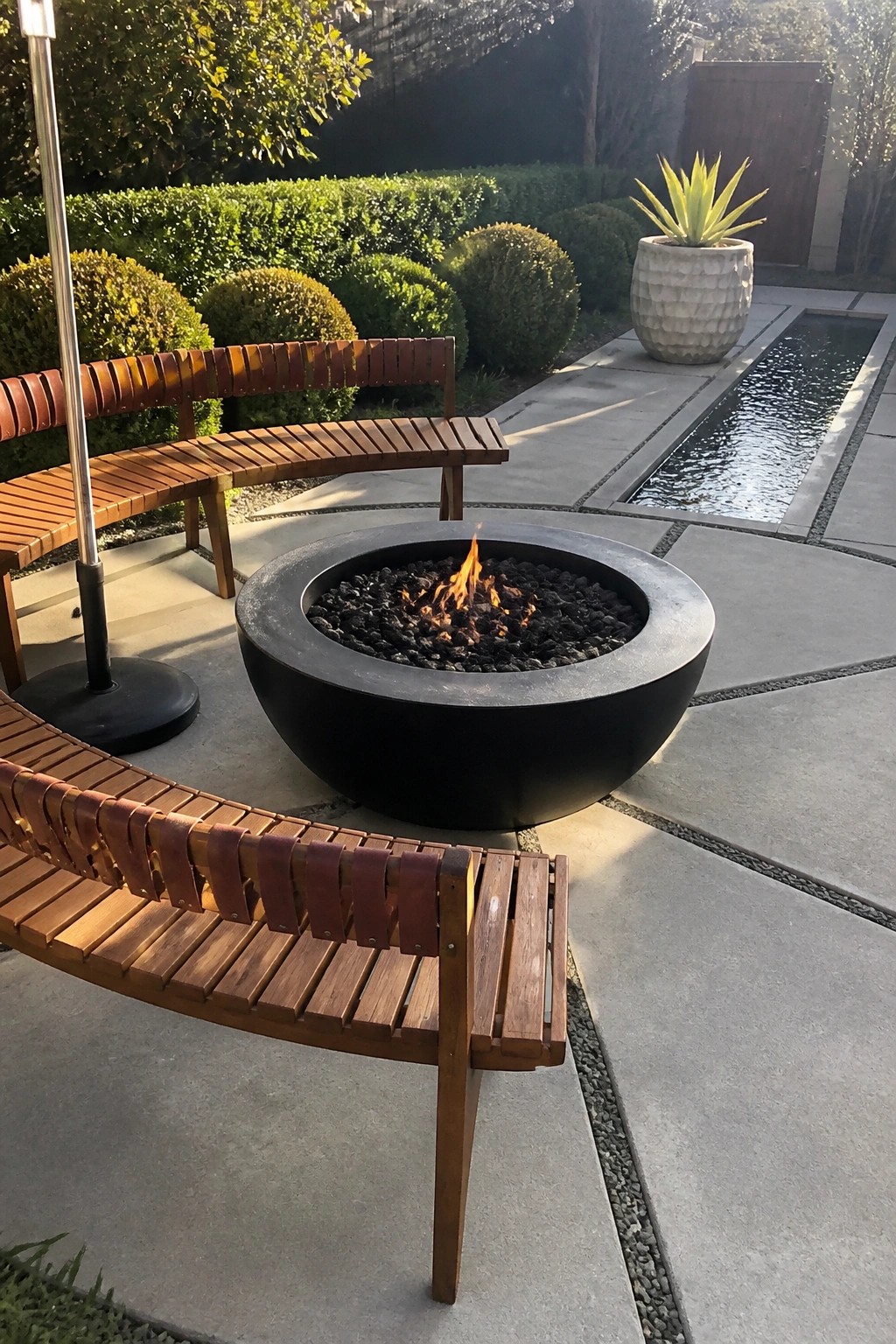 Curved wooden benches surround a flaming black fire pit on a stone patio with water feature.