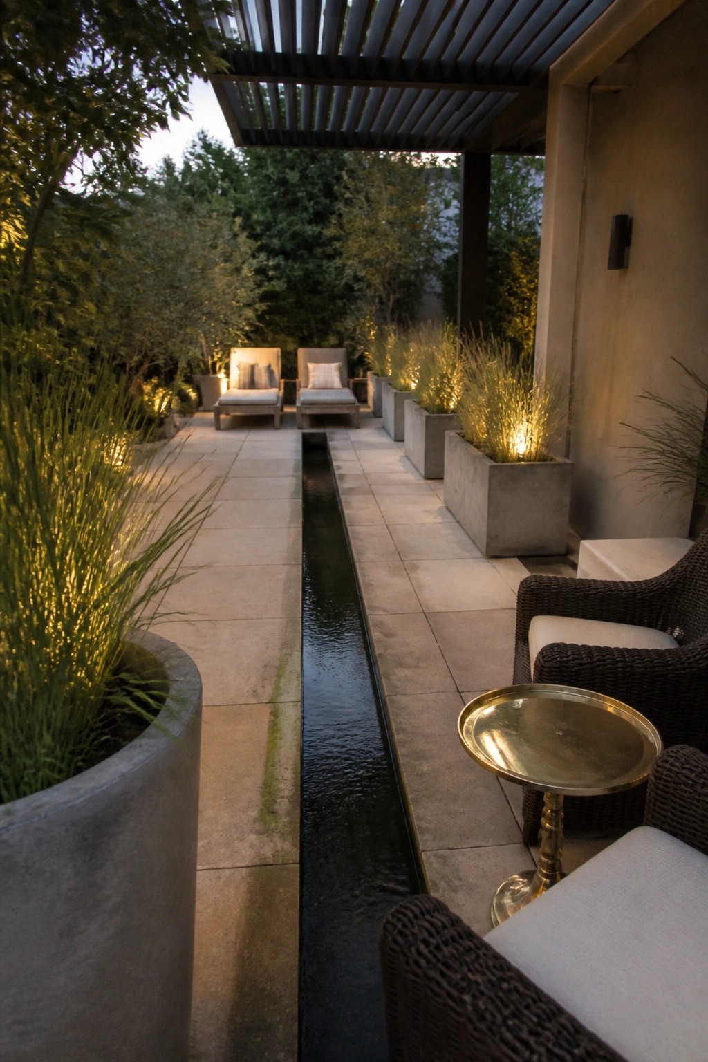 A narrow outdoor patio with a long central water channel, lounge chairs on both sides, and large concrete planters with grasses under a slatted pergola.