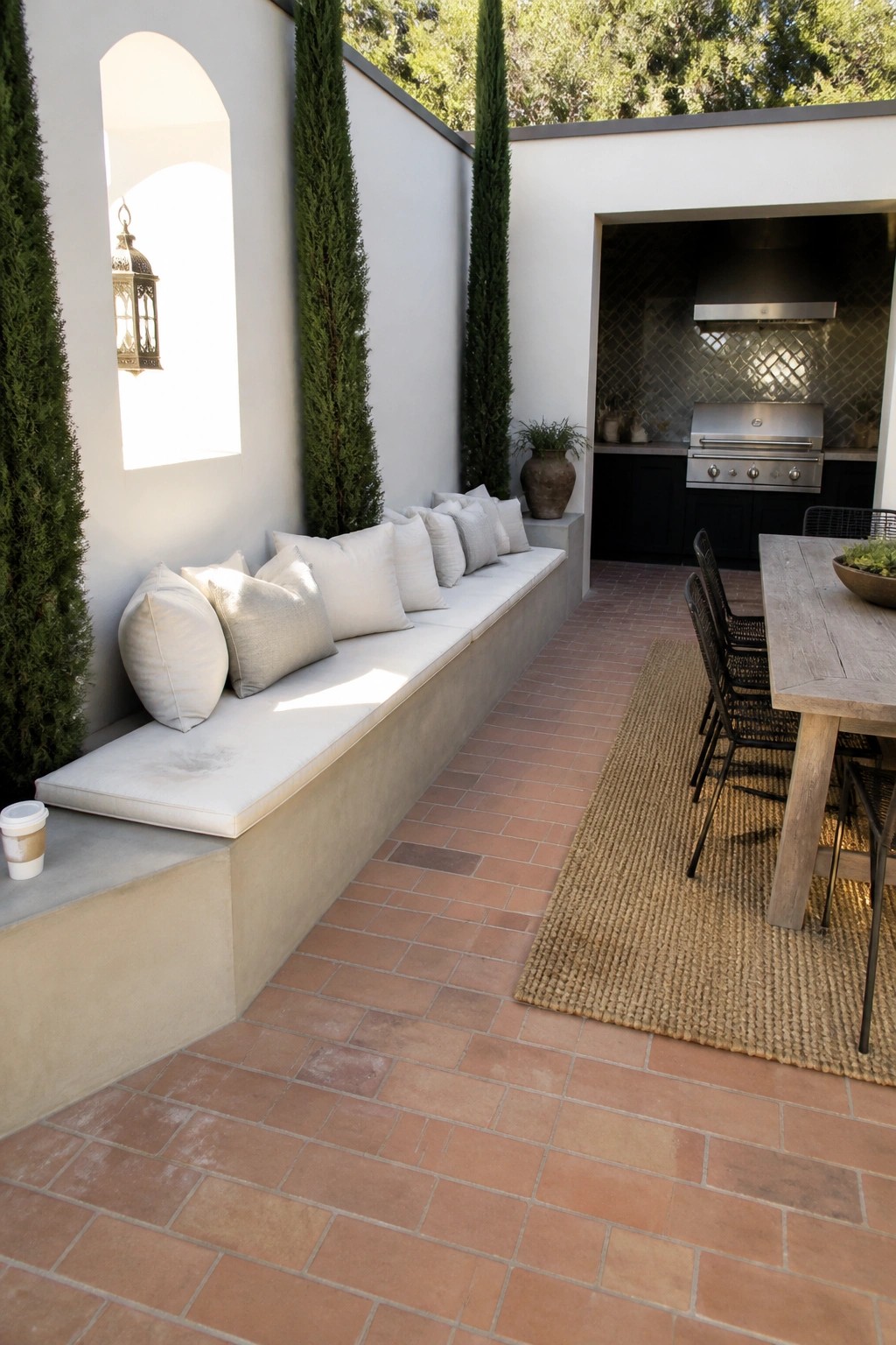 A narrow outdoor patio features a long built-in bench with white cushions along a white wall, a wooden dining table with chairs on a woven rug, and an outdoor kitchen visible in the background.