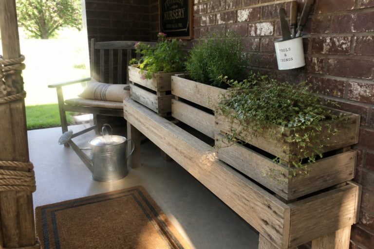 17 Rustic Pallet Planter Ideas Using Weathered and Natural Wood Wooden pallet bench with attached planters filled with green herbs and ivy on a brick porch beside a metal watering can.