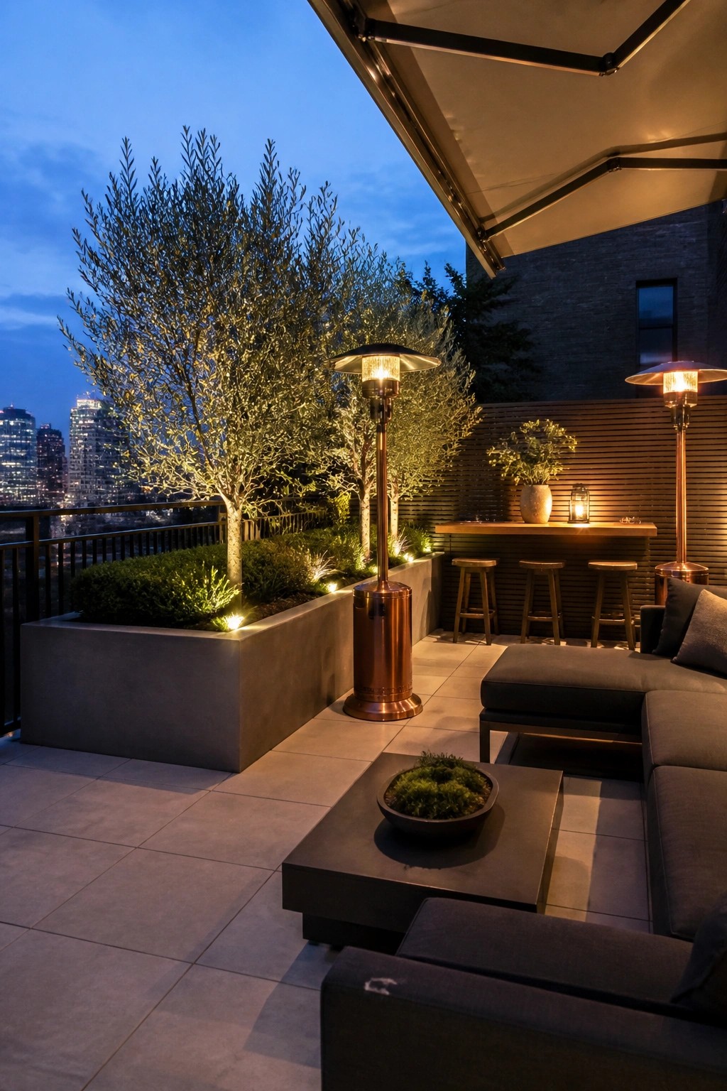 Modern rooftop patio at dusk with large concrete raised planter beds holding trees and shrubs, dark outdoor seating, a bar with stools, and two tall copper patio heaters overlooking a city skyline.