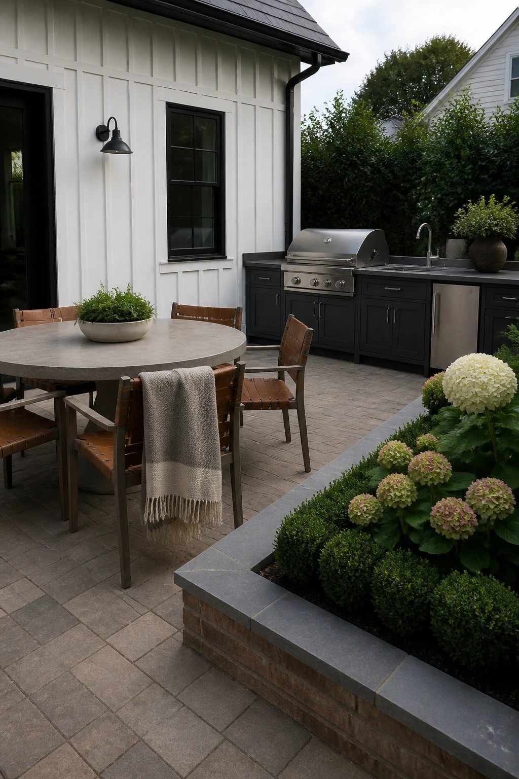 A paved patio with a round dining table and leather chairs beside a raised stone and brick border bed planted with green shrubs and hydrangeas, with a dark outdoor kitchen visible in the background.