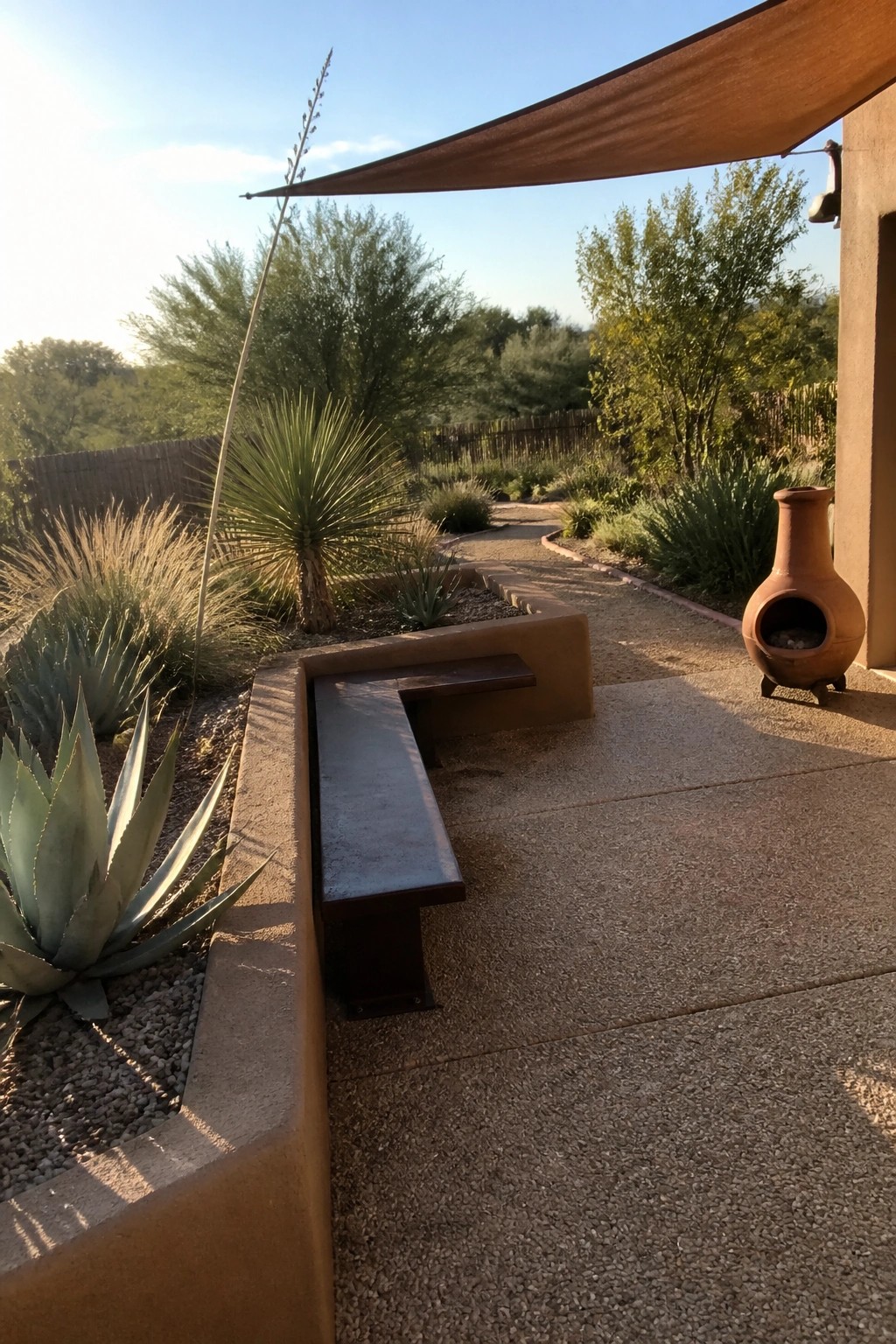 A concrete patio with built-in raised border beds containing agave and desert plants, a metal bench integrated into the bed, a gravel path, and a clay chiminea nearby.