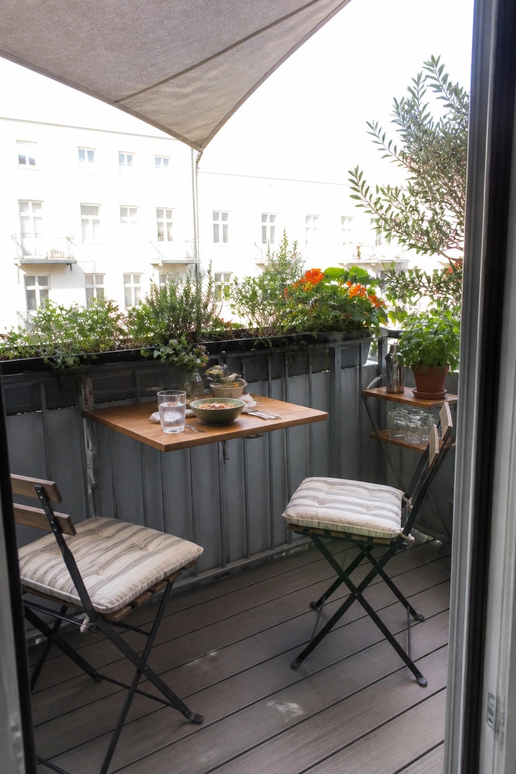 A compact balcony dining area with a wooden fold-down table attached to the railing, two folding chairs with striped cushions, and potted plants along the edge.