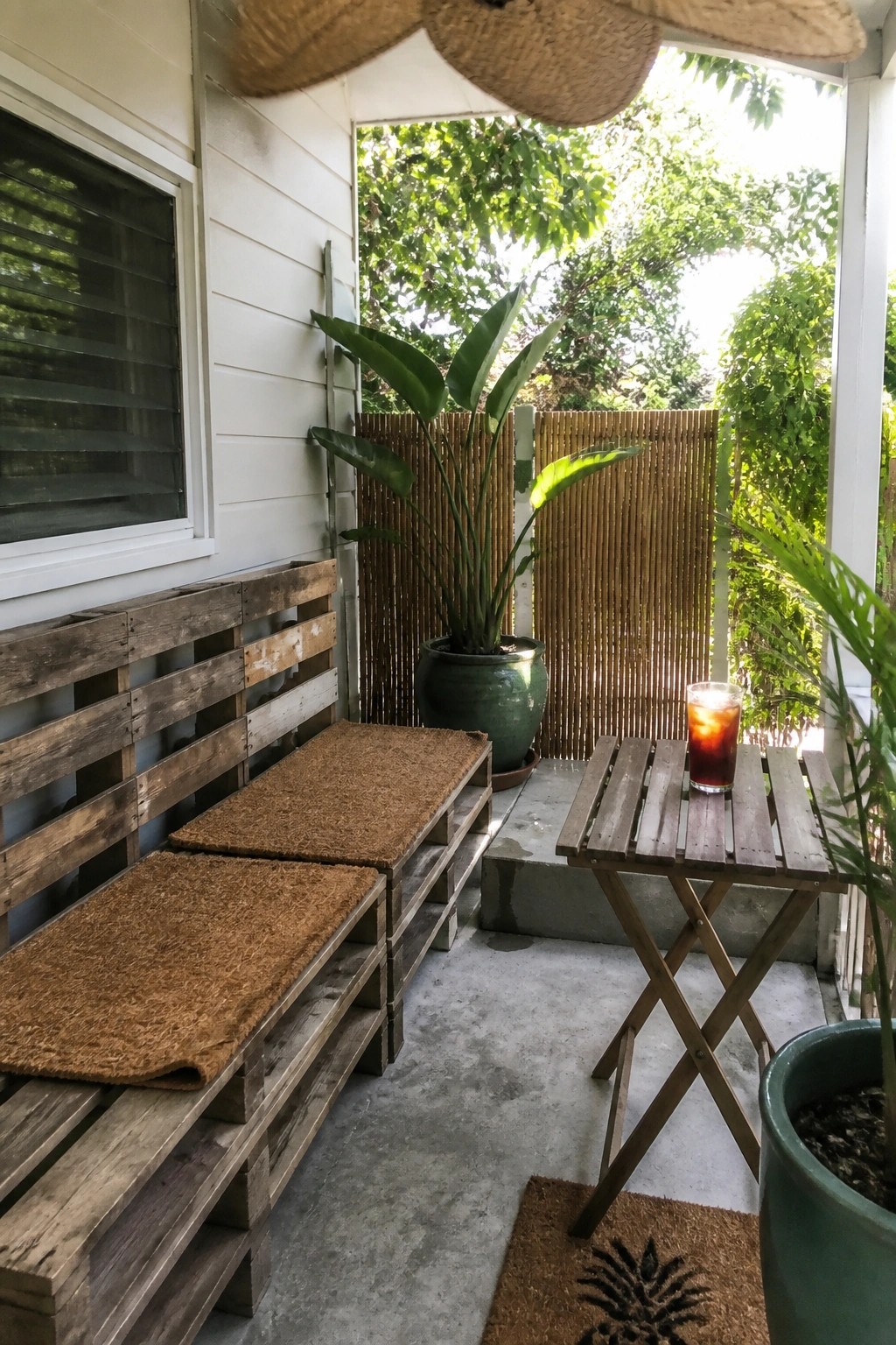 A narrow porch features wooden pallet benches with brown cushions along a white house wall, a small folding table with a glass of iced tea, and potted plants in front of a bamboo screen.