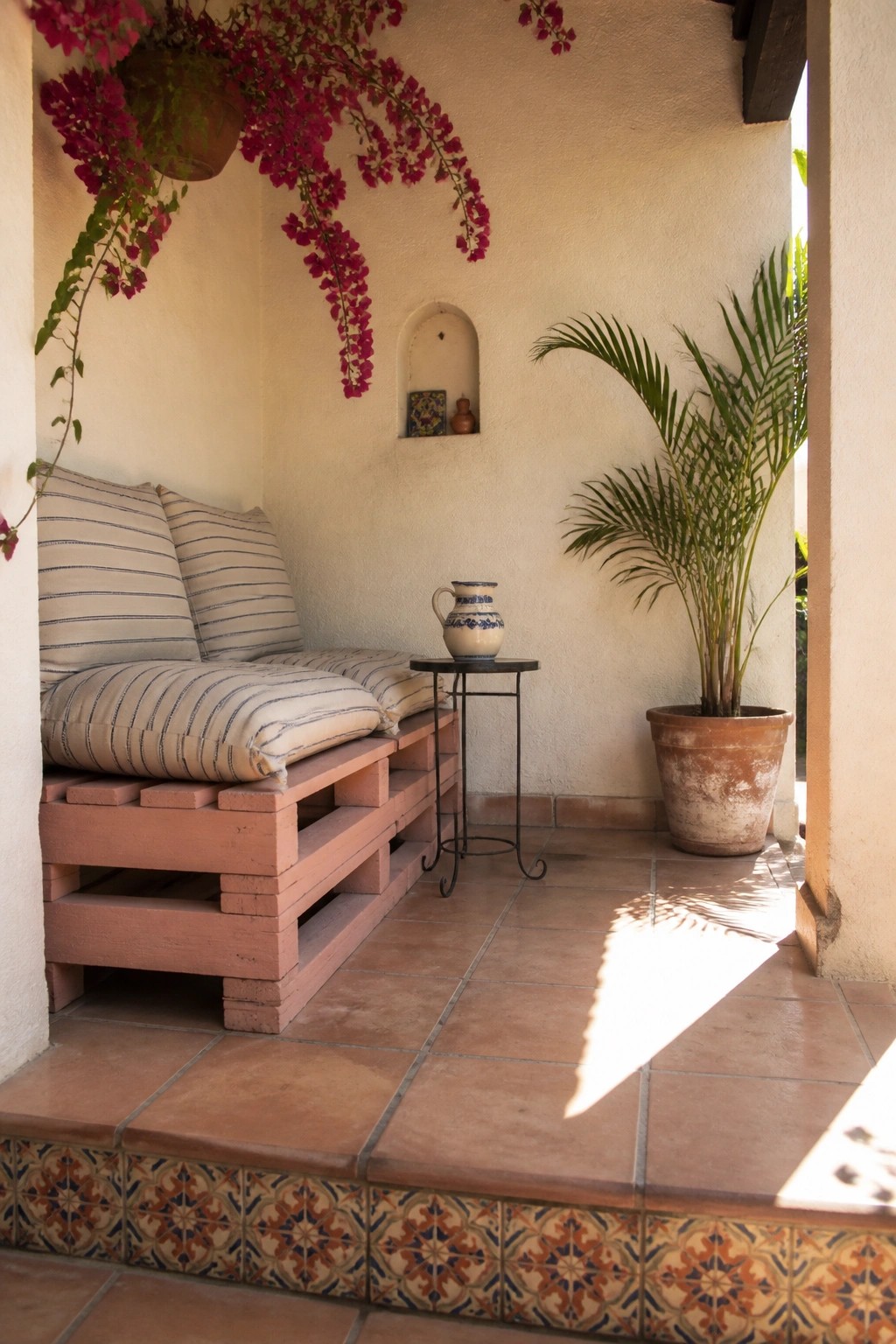 A painted wooden pallet bench with striped cushions on a tiled porch beside a potted palm tree.