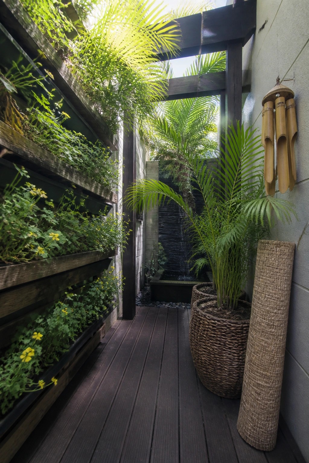 A narrow wooden deck path beside a tall vertical pallet planter filled with green plants and small yellow flowers, next to potted palms and a water feature.