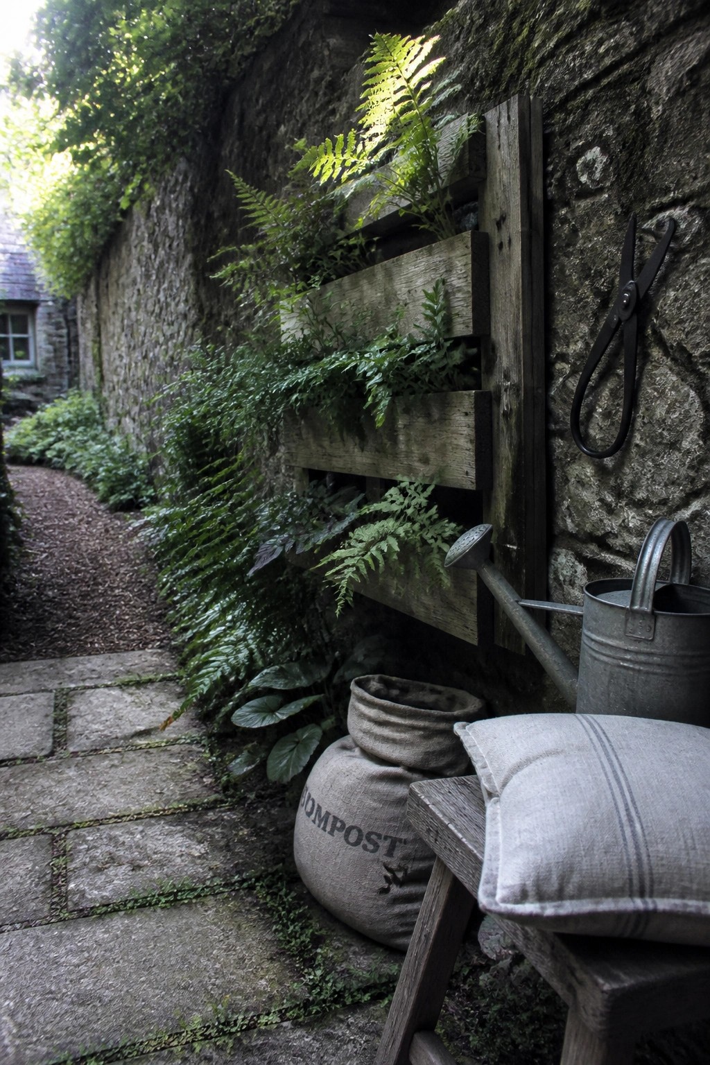 A wooden pallet mounted vertically on a stone wall holds multiple ferns, with a watering can, shears, and a compost sack on the ground nearby.