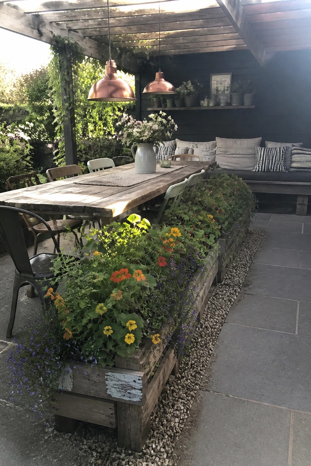A long wooden pallet planter filled with colorful cascading flowers runs alongside a rustic outdoor dining table on a stone patio.