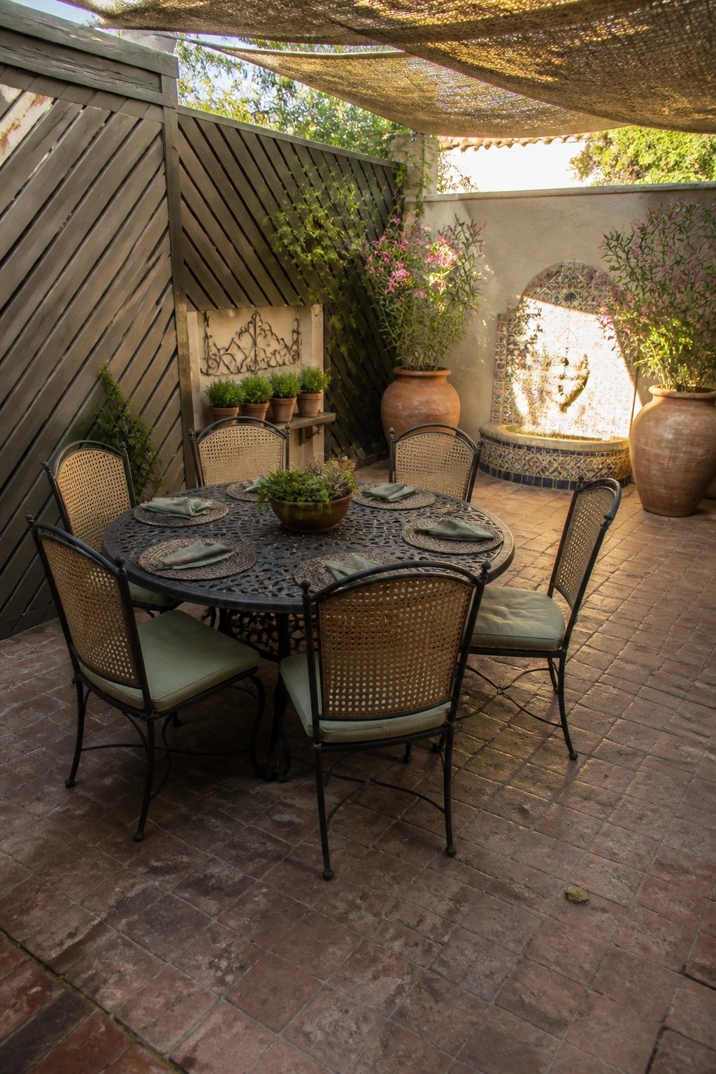 A round metal patio table with cane-back chairs sits on brick pavers beside a tall wooden slatted screen wall, with potted plants and a tiled fountain visible in the background.