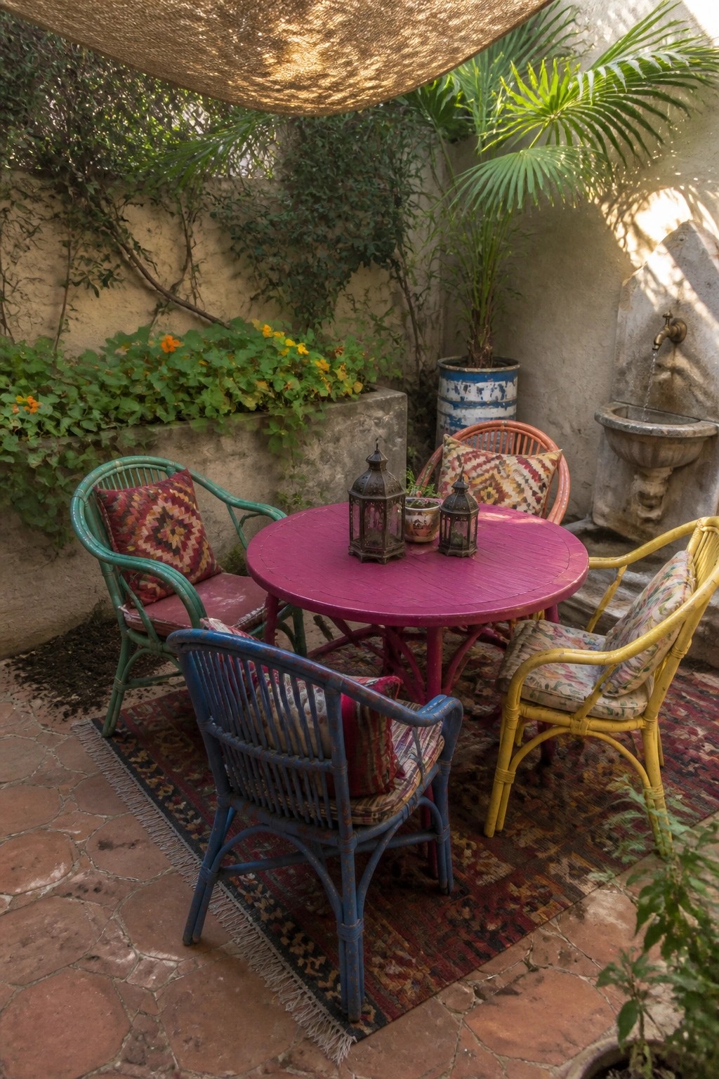 A patio scene with a round magenta table surrounded by four painted wicker chairs in green, blue, and yellow, set on a patterned rug with potted plants and a stone fountain nearby.