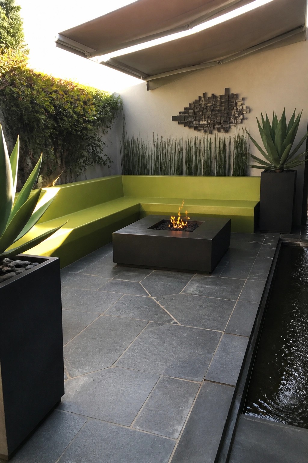 An outdoor patio with L-shaped lime green built-in seating around a square metal fire pit on gray stone tiles, next to potted agave plants and a narrow water channel.