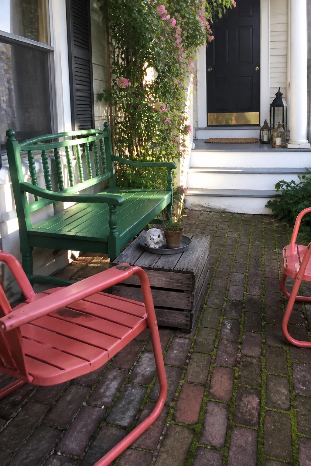 A patio scene with a green painted wooden bench, red metal chairs, a wooden crate table, and a black door in the background.