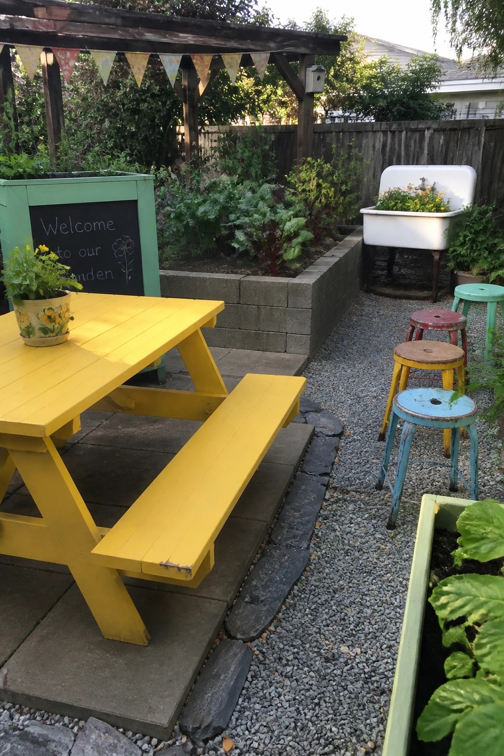 Yellow picnic table in colorful garden with chalkboard sign, stools, and raised beds